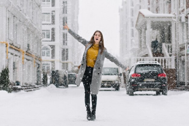 Anmutiges kaukasisches weibliches Modell im langen Mantel, der auf der Straße im Wintermorgen tanzt. Außenfoto der bezaubernden Dame im gelben Pullover, der Hände während des Fotoshootings am frostigen Tag winkt.