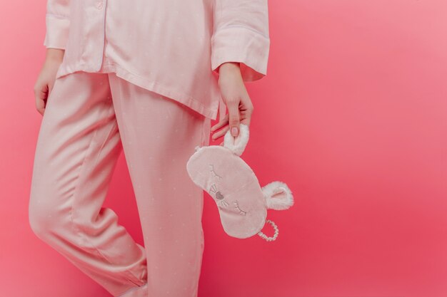 Anmutiges weibliches Modell im kuscheligen Nachtanzug, der Schlafmaske hält. Innenfoto der Frau trägt rosa Baumwollpyjamas, die mit Augenmaske in der Hand stehen.
