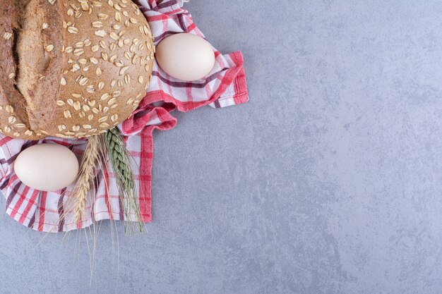 Anordnung von weizenstielen, eiern und einem laib brot auf einem handtuch auf marmoroberfläche