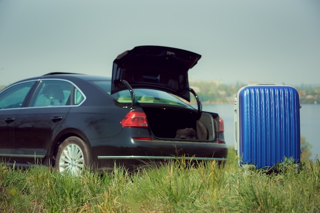 Ansicht des modernen schwarzen Autos und des blauen Koffers auf der Flussseite im sonnigen Tag.