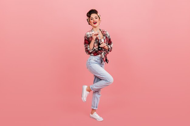 Ansicht des Pinup-Mädchens in voller Länge in Jeans. Studioaufnahme der lachenden eleganten Frau, die mit Sodaflasche aufwirft.