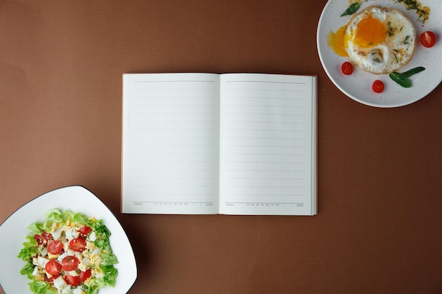 Ansicht von Notizblock und Spiegelei mit Tomaten-Dill in Teller mit Blättern und Gemüsesalat in Teller mit Tomaten-Ei-Salat auf braunem Hintergrund