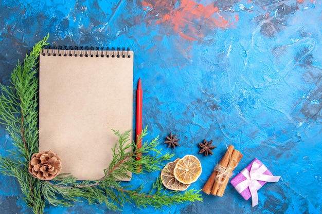 Ansicht von oben ein Notizbuch Pine Tree Branches Pinecone Red Pencil Zimtstangen Anis getrocknete Zitronenscheiben auf blauem Hintergrund Freiraum