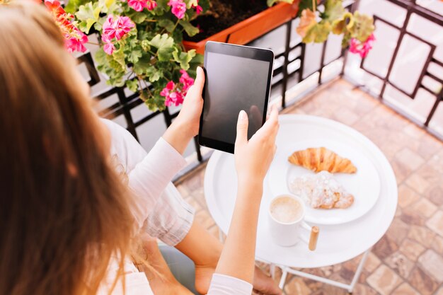 Ansicht von oben Tablette in den Händen des Mädchens im Pyjama, das Blumen auf Balkon sitzt und morgens frühstückt.