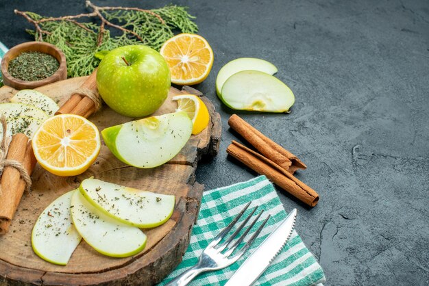 Ansicht von unten Apfelscheiben gebundene Zimtstangen und Zitronenscheiben Apfel mit Minze auf Holzbrett Kiefernzweigen eine Gabel und ein Messer auf grüner Serviette auf schwarzem Tisch mit Kopierplatz