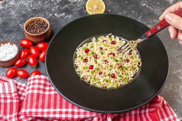 Ansicht von unten asiatische Ramen-Nudeln auf schwarzer Tellergabel in weiblicher Hand geschnittene Zitronen-Kirschtomaten Meersalz schwarzer Pfeffer in kleinen Schüsseln auf dunklem Tisch