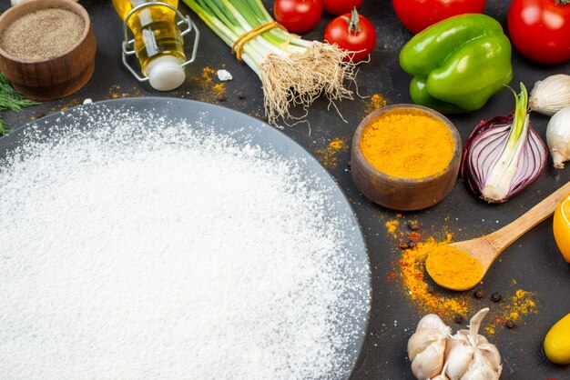 Ansicht von unten bestreutes Mehl auf runder Platte schwarzer Pfeffer und Kurkuma in kleinen Schalen Tomaten Knoblauch Frühlingszwiebeln auf Tisch