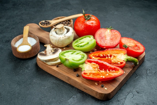 Ansicht von unten frisches Gemüse Pilze geschnitten rote und grüne Tomaten Paprika auf Schneidebrett Schalen mit schwarzem Pfeffer und Salz Holzlöffel auf dunklem Tisch