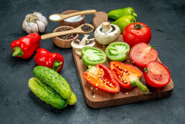 Ansicht von unten Gemüse Pilze rote und grüne Tomaten rote Paprika auf Schneidebrett Knoblauch schwarzer Pfeffer und Salz in Schalen Holzlöffel Gurken auf dunklem Tisch