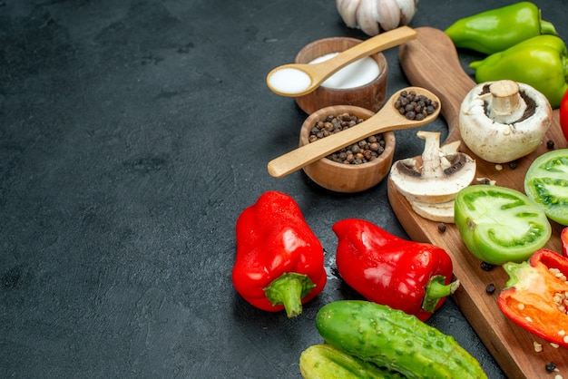 Ansicht von unten Gemüse Pilze Tomaten rote Paprika auf Schneidebrett Knoblauch schwarzer Pfeffer und Salz in Schalen Holzlöffel Gurken auf dunklem Tisch mit Kopie Platz