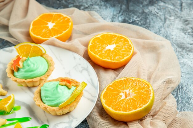 Ansicht von unten kleine Torten mit grüner Gebäckcreme und Zitronenscheibe auf Teller auf beigem Schal geschnittene Orangen auf dunkler Oberfläche
