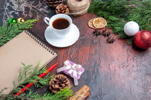 Ansicht von unten roter Stift ein Notizbuch Kiefer Zweige Weihnachtsbaum Ball Spielzeug und Geschenk Zimt Anis Stroh Faden eine Tasse Tee auf dunkelrotem Hintergrund freier Raum