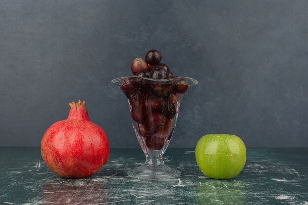 Apfel, Granatapfel und Glas schwarze Trauben auf Marmortisch.