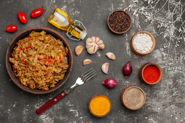 appetitliches Gericht grüne Bohnen und Tomaten neben der Gabel Flasche Öl Knoblauch Zwiebel Tomaten und Schüsseln mit bunten Gewürzen auf dem dunklen Tisch