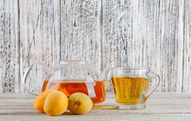 Aprikosentee in der Teekanne und im Glasbecher mit Seitenansicht der Aprikosen auf einem Holztisch