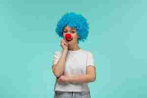 Kostenloses Foto aprilscherz lächelndes mädchen, das denkt, ellbogen mit roter nase in einem blauen haar des clownkostüms zu berühren