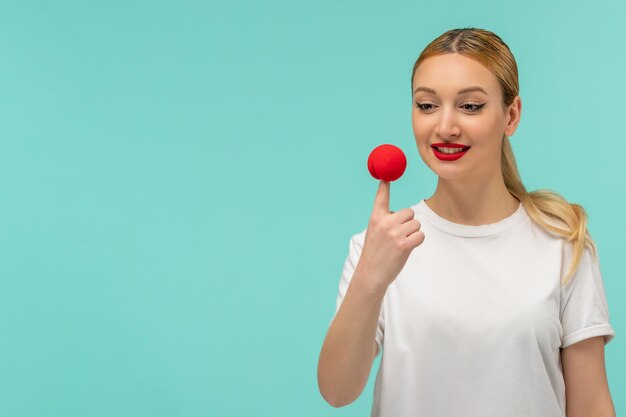 Aprilscherztag blondes lächelndes Mädchen Pferdeschwanz rote Nase auf Finger weißes T-Shirt Clown lustig