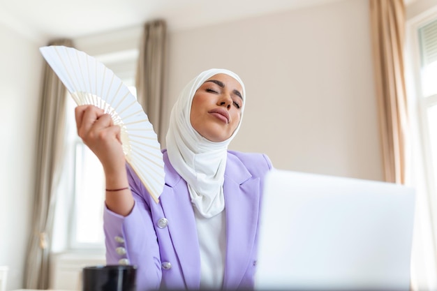 Arabische Büroangestellte, die unter Hitze, heißer, stickiger Luft leiden und an Sommertagen von zu Hause aus ohne Klimaanlage am Laptop arbeiten Muslimische Geschäftsfrau kühlt mit Handventilator am Arbeitsplatz ab