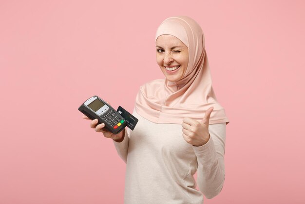 Arabische muslimische frau in hijab-leichter kleidung, die isoliert auf rosafarbenem hintergrund posiert. menschen religiöses lebensstilkonzept. mock-up-kopienbereich. zahlungsterminal zur verarbeitung halten, kreditkartenzahlungen erwerben.