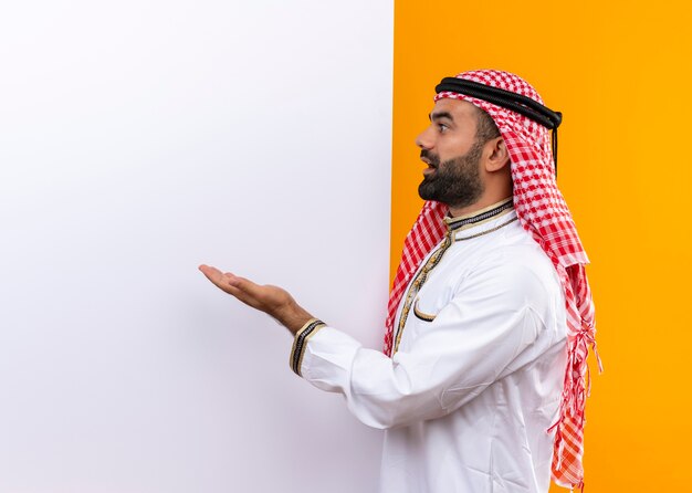 Arabischer Geschäftsmann in der traditionellen Abnutzung, die auf leere Plakatwand mit Arm zeigt, der überrascht steht über orange Wand steht