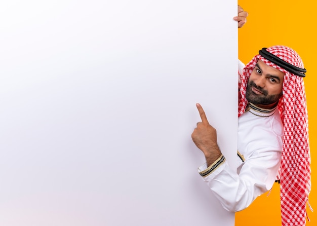 Arabischer Geschäftsmann in der traditionellen Abnutzung, die leere Plakatwand herausspäht, die mit dem Finger darauf lächelt und über orange Wand steht