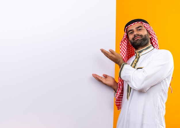 Arabischer Geschäftsmann in der traditionellen Abnutzung, die mit der leeren Plakatwand der Arme über orange Wand steht