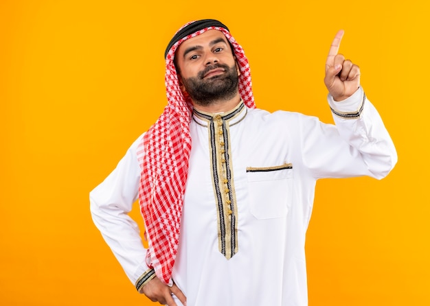 Arabischer Geschäftsmann in der traditionellen Abnutzung, die zuversichtlich zeigt, mit dem Finger zur Seite stehend über orange Wand zu zeigen