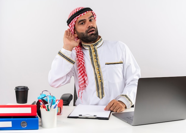 Arabischer Geschäftsmann in der traditionellen Kleidung, die am Tisch mit Laptop-Computer hält Hand in der Nähe seines Ohrs sitzt und versucht, im Büro zuzuhören