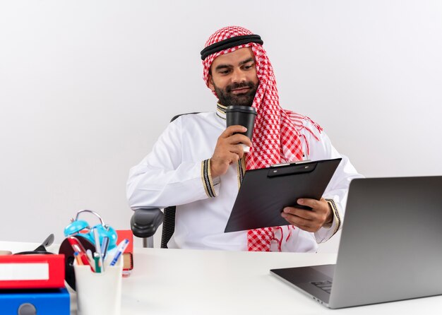 Arabischer Geschäftsmann in der traditionellen Kleidung, die am Tisch mit Laptop-Computer hält Zwischenablage und Kaffeetasse glücklich und positiv im Büro arbeitet