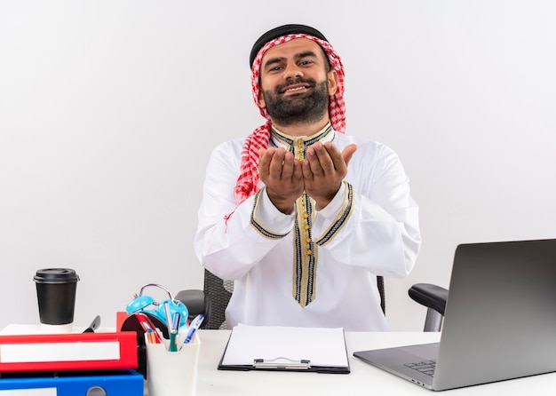 Arabischer Geschäftsmann in der traditionellen Kleidung, die am Tisch mit Laptop-Computer sitzt, der Hände zusammenhält, die um Geld bitten, das im Büro arbeitet