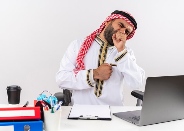 Arabischer Geschäftsmann in der traditionellen Kleidung, die am Tisch mit Laptop-Computer sitzt, der müde gähnt, das im Büro arbeitet