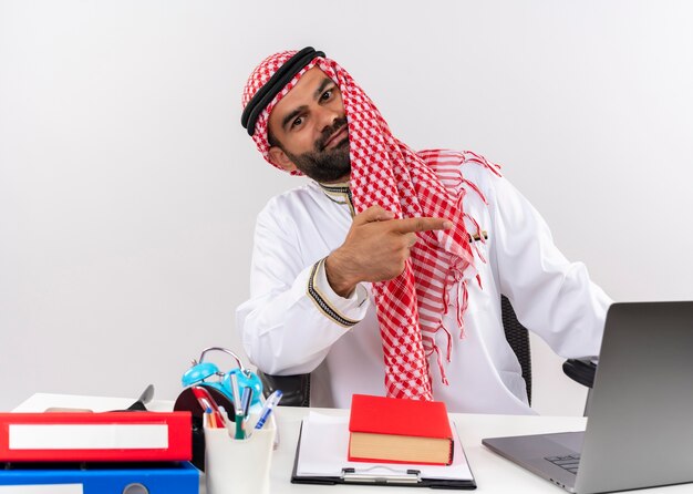 Arabischer Geschäftsmann in der traditionellen Kleidung, die am Tisch mit Laptop-Computern sitzt, die sicher zeigen, mit dem Finger zur Seite zu zeigen, die im Büro arbeitet