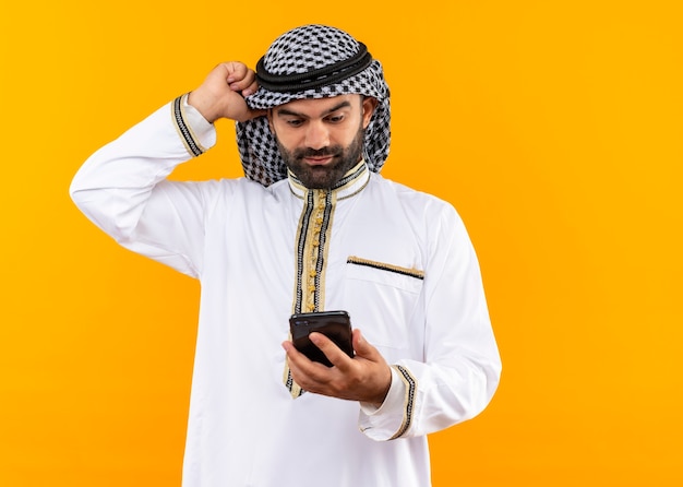 Arabischer Geschäftsmann in der traditionellen Kleidung, die Bildschirm seines Smartphones verwirrt betrachtet, der über orange Wand steht