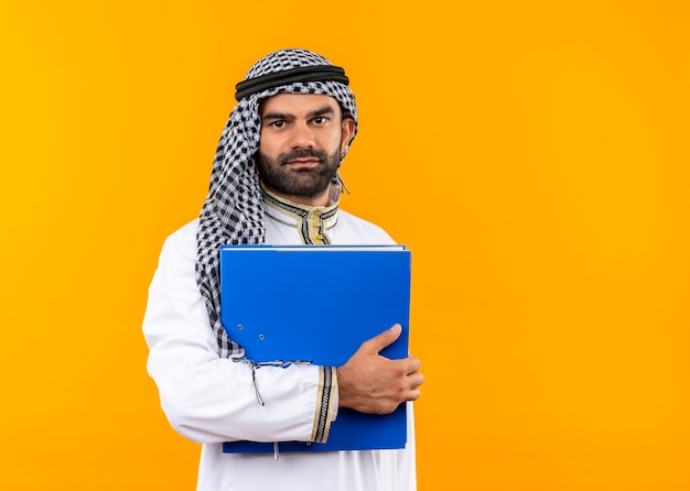 Arabischer Geschäftsmann in der traditionellen Kleidung, die blauen Ordner hält, der zuversichtlich steht, über orange Wand zu stehen