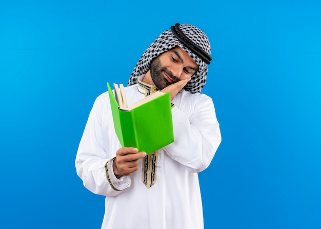 Arabischer Geschäftsmann in der traditionellen Kleidung, die ein offenes Buch hält, das es mit Lächeln auf Gesicht betrachtet, das über blauer Wand steht