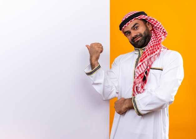Arabischer Geschäftsmann in der traditionellen Kleidung, die nahe der leeren Plakatwand steht, die mit dem Finger darauf mit dem selbstbewussten Lächeln auf Gesicht über orange Wand zeigt