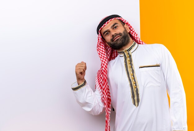 Arabischer Geschäftsmann in der traditionellen Kleidung, die nahe der leeren Plakatwand steht, die mit dem Finger darauf mit dem selbstbewussten Lächeln auf Gesicht über orange Wand zeigt