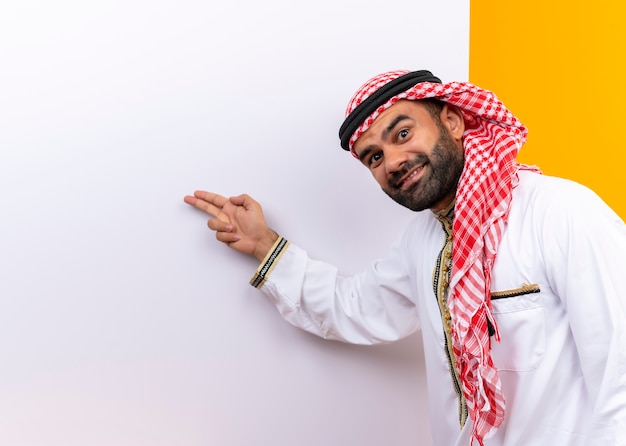 Arabischer Geschäftsmann in der traditionellen Kleidung, die nahe der leeren Plakatwand steht, die mit den Fingern darauf mit dem Lächeln auf Gesicht über orange Wand zeigt