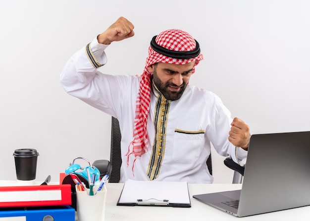 Arabischer Geschäftsmann in traditioneller Kleidung, die am Tisch mit geballten Fäusten des Laptop-Computers mit wütendem Gesicht sitzt, das im Büro arbeitet