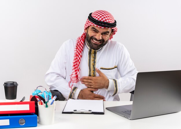 Arabischer Geschäftsmann in traditioneller Kleidung, die am Tisch mit Laptop-Computer lacht und seinen Bauch berührt, der im Büro arbeitet