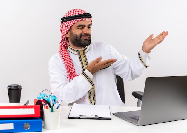 Arabischer Geschäftsmann in traditioneller Kleidung, die am Tisch mit Laptop-Computer sitzt, der mit Armen zur Seite zeigt, die im Büro arbeiten