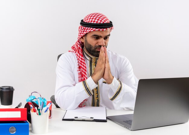 Arabischer Geschäftsmann in traditioneller Kleidung, die mit Laptop-Computer arbeitet, der Hände zusammen mit Hoffnungsausdruck hält, der am Tisch im Büro sitzt