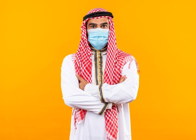 Arabischer Geschäftsmann in traditioneller Kleidung und Gesichtsschutzmaske mit sicherem Ausdruck, der über orange Wand steht