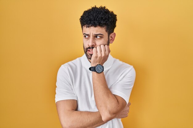 Arabischer Mann, der über gelbem Hintergrund steht und gestresst und nervös aussieht, mit den Händen auf dem Mund, die Nägel beißen. Angstproblem.