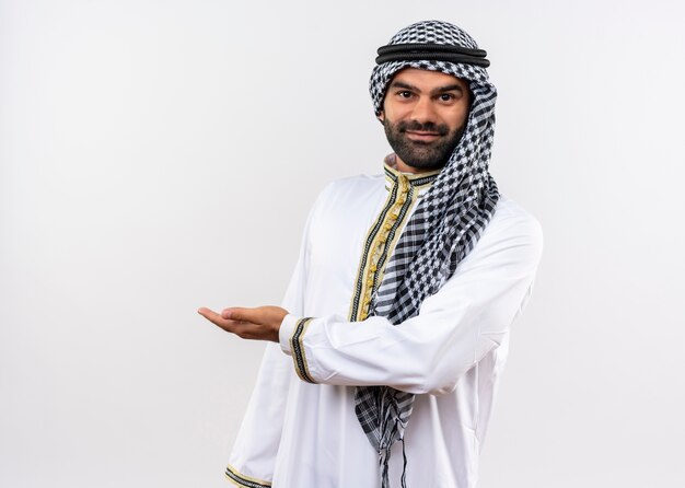 Arabischer Mann in der traditionellen Kleidung, die lächelnd mit Arm der Hand steht, die über weißer Wand steht