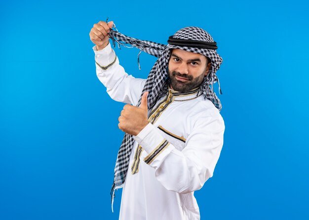 Arabischer Mann in der traditionellen Kleidung lächelnd, die Daumen oben stehend über blaue Wand zeigt