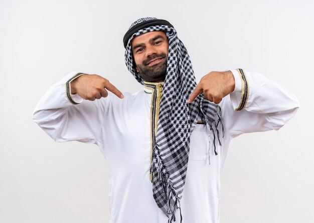 Arabischer Mann in traditioneller Kleidung, der mit den Fingern auf sich selbst zeigt, selbstzufrieden und stolz, über weißer Wand stehend