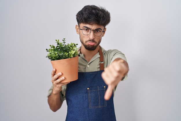 Kostenloses Foto arabischer mann mit bart, der einen grünen blumentopf in der hand hält, sieht unglücklich und wütend aus und zeigt ablehnung und negativität mit gesenktem daumen und gestikuliert mit schlechtem gesichtsausdruck