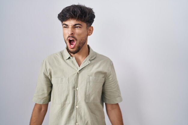 Arabischer Mann mit Bart, der über weißem Hintergrund steht, wütend und wütend, schreit frustriert und wütend und schreit vor Wut. Wut und aggressives Konzept.