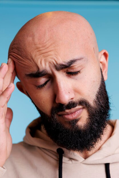 Arabischer Mann mit geschlossenen Augen, der Migräne hat und sich vor Schmerzen den Kopf reibt. Junger Erwachsener mit kahlem Bart, der unter Kopfschmerzen leidet und die Schläfe hält, während er auf blauem Hintergrund posiert